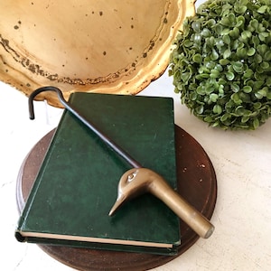 May include: A metal fireplace tool featuring a handle shaped like a duck head in a brass finish, connected to a long, slender, dark-colored iron rod that curves into a hook at the top end, resting diagonally across a dark green hardcover book.