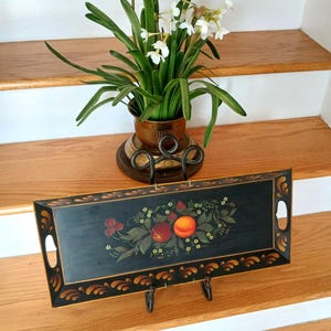 May include: A rectangular black wooden serving tray with a painted fruit design, including peaches and berries, and decorative gold trim. The tray is displayed on a black metal stand. A potted plant with white flowers sits behind the tray.