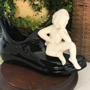 May include: A black ceramic shoe-shaped planter with a glossy finish. A white figurine of a seated child is perched on the shoe. The planter sits on a dark wooden base, with greenery in the background.