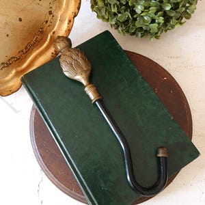 May include: A wall hook featuring a brass finial shaped like a pineapple or artichoke, attached to a curved black metal hook. The decorative brass top has detailed leaf-like textures, while the lower section is a smooth, solid black metal J-hook.