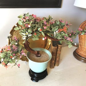 May include: A decorative bonsai tree with pink and white flowers, green leaves, and small red and white berries. The tree is in a light blue pot on a black stand. A gold-colored decorative plate and two old books are in the background.