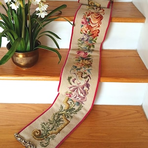 May include: A long, narrow tapestry runner featuring a beige woven background with a central vertical floral and human figure design in shades of red, pink, purple, and green. The runner has a red border and ornate brass hardware at both ends.