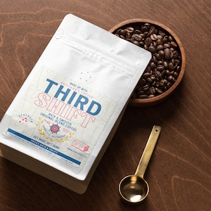 Puede incluir: Una bolsa de café blanca con el texto "THIRD SHIFT" en azul y rojo, y "ORIGINAL BLEND COFFEE" impreso en ella. Un cuenco de madera con granos de café y una cuchara medidora dorada están junto a la bolsa. La bolsa contiene 113g de café.