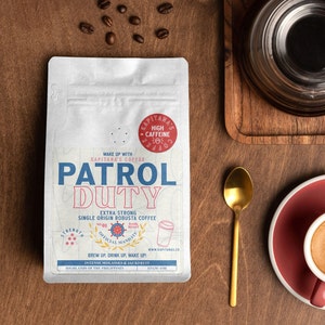 Op de afbeelding: Een witte koffiezak met een rood en blauw logo waarop "Patrol Duty" en "Extra Strong Single Origin Robusta Coffee" staat. De zak ligt op een houten oppervlak met een kopje koffie en een lepel.