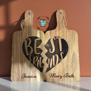 May include: Two wooden cutting boards with a heart design that reads "Best Friends" when placed together. One board is labeled "Jessica" and the other "Mary Beth". Each board has a handle with a jute string.