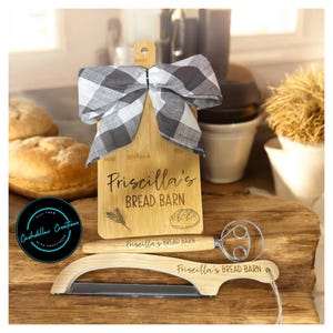 May include: A wooden bread cutting board with a black and white checkered bow, a bread lame, and a wire whisk. The cutting board is engraved with "Priscilla's BREAD BARN". A round logo is in the lower left corner.