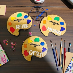May include: Three wooden artist palettes with colorful paint splotches and the names "Nathan", "Mackenzie", and "Carson". Each palette has the words "Artwork By" above the names. Paintbrushes and art supplies are visible.