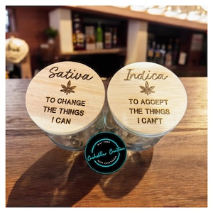 May include: Two wooden lids for glass jars with engraved text and leaf designs. The left lid says "Sativa" with the text "To change the things I can". The right lid says "Indica" with the text "To accept the things I can't".