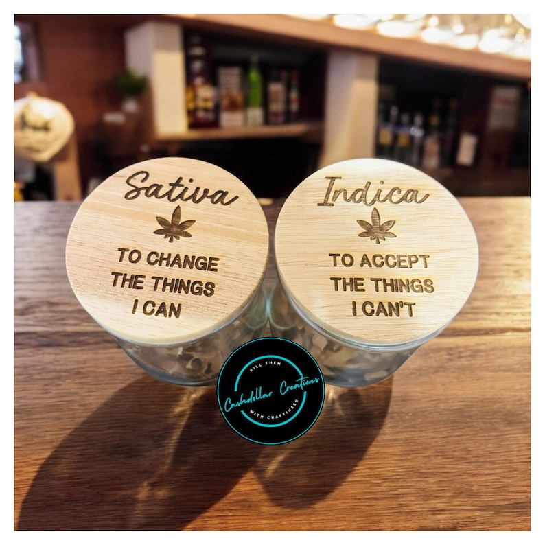 May include: Two wooden lids for glass jars with engraved text and leaf designs. The left lid says "Sativa" with the text "To change the things I can". The right lid says "Indica" with the text "To accept the things I can't".