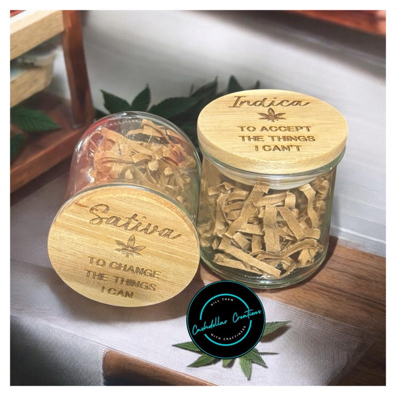 May include: Two glass jars with wooden lids. The lids are engraved with the words "Sativa" and "Indica" and a marijuana leaf. The Sativa lid reads "To change the things I can". The Indica lid reads "To accept the things I can't".