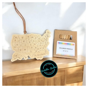 May include: Wooden US map ornament with a brown leather cord for hanging. The map is outlined with state borders and includes the text "Oh, the places we go." A box of 12 colored pencils is included.