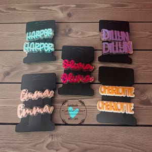 May include: Several personalized hair clips with names like "Harper," "Dillyn," "Olivia," "Emma," and "Cheriotte" are displayed on black cards. The clips are in various colors, including blue, purple, pink, and orange, with glitter accents. The words "Handmade" and "with care" are also visible.