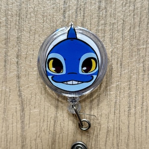 K&ouml;nnte beinhalten: Ein einziehbarer Ausweis-Clip mit einem Cartoon-Hai-Gesicht. Das Design zeigt einen blauen Hai mit wei&szlig;en und gelben Details, umh&uuml;llt von klarem Kunststoff. Er hat einen Metallclip zum Befestigen an der Kleidung.