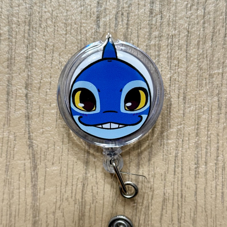 K&ouml;nnte beinhalten: Ein einziehbarer Ausweis-Clip mit einem Cartoon-Hai-Gesicht. Das Design zeigt einen blauen Hai mit wei&szlig;en und gelben Details, umh&uuml;llt von klarem Kunststoff. Er hat einen Metallclip zum Befestigen an der Kleidung.