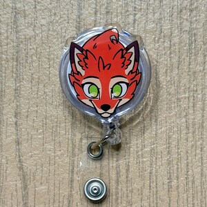 K&ouml;nnte beinhalten: Ein durchsichtiges, einziehbares Ausweis-Jojo mit einem Cartoon-Fuchsgesicht. Der Fuchs hat rotes Fell, schwarze Akzente und gr&uuml;ne Augen. Die Rolle hat einen silbernen Metallclip und einen transparenten Kunststoff-Druckknopf.