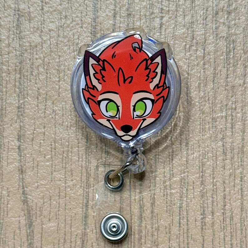 K&ouml;nnte beinhalten: Ein durchsichtiges, einziehbares Ausweis-Jojo mit einem Cartoon-Fuchsgesicht. Der Fuchs hat rotes Fell, schwarze Akzente und gr&uuml;ne Augen. Die Rolle hat einen silbernen Metallclip und einen transparenten Kunststoff-Druckknopf.