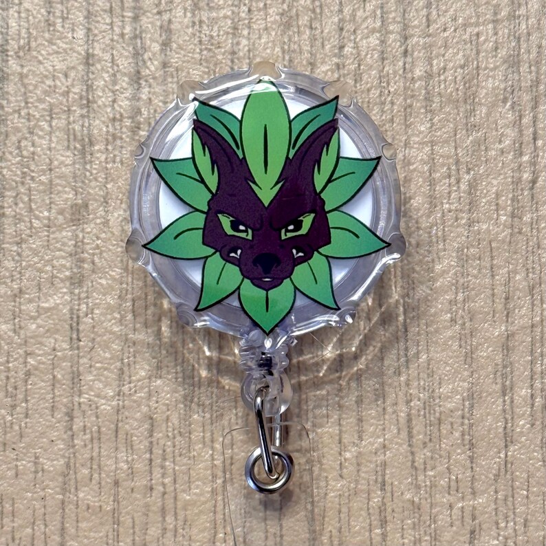 K&ouml;nnte beinhalten: Ein klarer, runder, einziehbarer Ausweishalter mit einem stilisierten lila und gr&uuml;nen Wolfsgesicht-Design. Der Wolf ist von gr&uuml;nen Bl&auml;ttern umgeben. Der Ausweishalter hat einen silbernen Metallclip und eine transparente Kunststoffschnur.