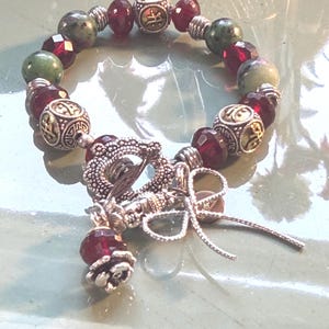 May include: A beaded bracelet featuring a mix of green, burgundy, and silver beads. The bracelet includes decorative silver beads with an ornate design and a silver clasp with a dangling charm. The beads are strung on a flexible cord.