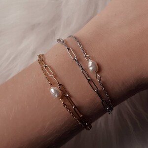 Op de afbeelding: Twee gouden en zilveren kettingarmbanden met parel accenten. De armbanden zijn op elkaar gelegd op iemands pols.