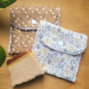 May include: Two fabric pouches with white buttons and floral patterns. The pouches are made of brown and white fabric with a white fringe around the edges. A bar of soap is sitting on top of the pouches.