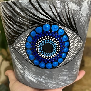 May include: Gray painted flower pot with a large eye design. The eye features a blue iris with white and blue dot details, surrounded by a gray and white painted background. Black lines create the eyelashes.