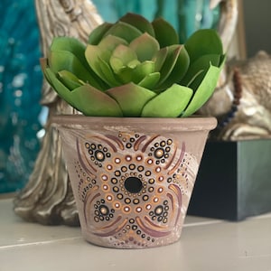 May include: A hand-painted terracotta plant pot with a geometric pattern in brown, gold, and black. It holds a green succulent. The pot is displayed on a white surface.