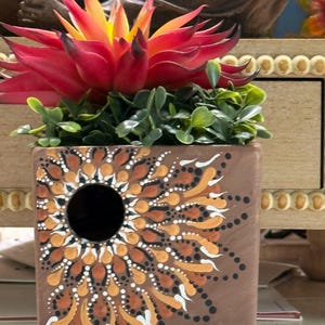 May include: A square brown ceramic planter featuring a hand-painted mandala design in white, gold, and dark brown dots. A vibrant red and yellow artificial succulent plant sits inside the planter, which has a circular opening on the front face.