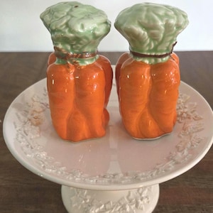May include: A pair of ceramic salt and pepper shakers shaped like carrots, with orange bodies and green tops. They are displayed on a white, footed cake stand with a floral design. The shakers have a glossy finish.