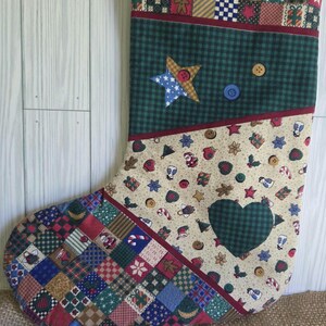 May include: A quilted Christmas stocking featuring a patchwork top border, a dark green section with a star and buttons, a cream-colored middle section with holiday icons, and a patchwork toe. A dark green heart is appliqued on the cream section.