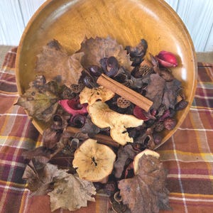 May include: A wooden bowl filled with dried leaves, apple slices, cinnamon sticks, and other potpourri. The bowl is light brown and sits on a plaid surface. The potpourri includes shades of brown, red, and beige, creating a warm, autumnal aesthetic.