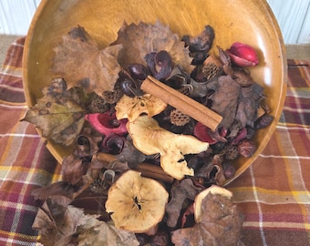 May include: A wooden bowl filled with dried leaves, apple slices, cinnamon sticks, and other potpourri. The bowl is light brown and sits on a plaid surface. The potpourri includes shades of brown, red, and beige, creating a warm, autumnal aesthetic.