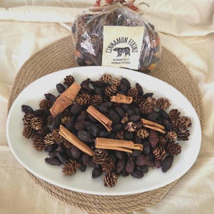 May include: A white oval platter filled with potpourri, including small pine cones, dark berries, and cinnamon sticks. A clear bag of potpourri tied with a plaid ribbon sits behind the platter. The bag has a label that reads "Cinnamon Fixins".