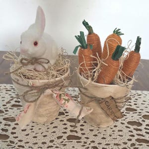 May include: Two decorative Easter-themed pots. One pot features a white bunny with pink-lined ears nestled in straw. The other pot contains several orange carrots with green tops. Both pots are tied with twine and adorned with floral fabric.