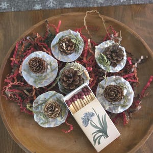 May include: A wooden bowl holds six decorative ornaments, each featuring a small pine cone on a light blue base with greenery. A matchbox with a floral design sits nearby. The bowl is filled with red and brown shredded paper.