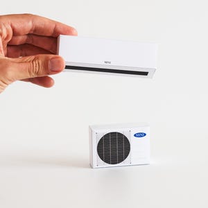 Op de afbeelding: Twee witte miniatuur airconditioning units. Eén wordt door een hand vastgehouden en heeft een zwarte ventilatieopening en het woord "MINI". De andere heeft een cirkelvormige ventilator en een blauw logo. De achtergrond is wit.