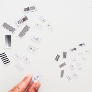 May include: A collection of small, white stickers with black graphic designs, including electrical outlets, light switches, and striped patterns. The stickers are scattered on a white surface. A hand is holding one of the outlet stickers.