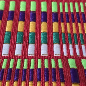 May include: Close-up of a red fabric with a repeating pattern of vertical columns. The columns are composed of stacked blocks in various colors, including green, purple, yellow, and white. The fabric has a textured appearance.