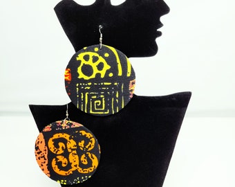 May include: A pair of round, black fabric earrings with colorful African print designs. One earring features a yellow and orange pattern, while the other has a red and orange pattern.