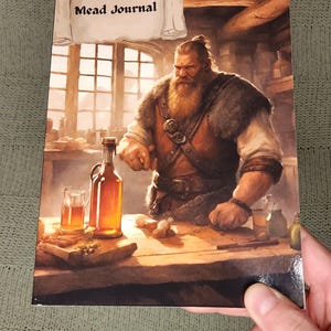May include: A mead journal cover featuring a bearded man in a brown leather vest and a white shirt, holding a bottle of mead. The journal cover has the text "Mead Journal" on a scroll.
