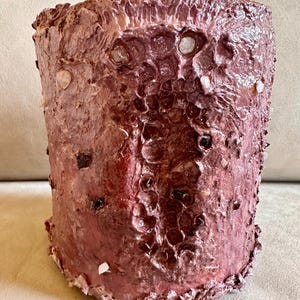 May include: A cylindrical, textured art piece in shades of burgundy and mauve. The surface has a rough, organic appearance with irregular patterns and embedded, light-colored accents. The overall aesthetic suggests a handcrafted, unique decorative item.