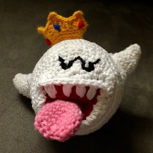 Puede incluir: Un juguete de peluche King Boo tejido a crochet con una corona amarilla, un cuerpo blanco, ojos negros, una boca roja y una lengua rosa.