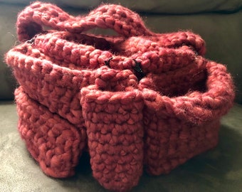 Puede incluir: Un bolso hecho a mano, tejido a ganchillo, en color rosa apagado. El bolso presenta una puntada texturizada y gruesa y un diseño estructurado con múltiples compartimentos y un asa. El bolso se asegura con cierres negros.