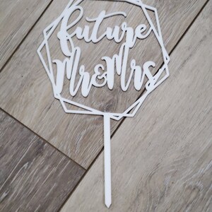 Può includere: Un cake topper bianco con le parole "future Mr & Mrs" in un carattere a script. Il testo è all'interno di una cornice geometrica a più lati. Il topper ha un lungo e sottile stelo per l'inserimento in una torta.