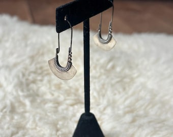 Puede incluir: Un par de pendientes plateados exhibidos en un soporte negro. Cada pendiente presenta un diseño curvo en forma de abanico con un borde texturizado y un gancho colgante. Se presentan sobre un fondo blanco suave.