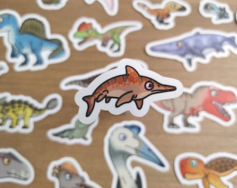 May include: A collection of dinosaur stickers, featuring various species in cartoon style. The stickers include a blue Spinosaurus, a green and red dinosaur, and a brown and orange fish-like dinosaur. The stickers are on a light brown surface.