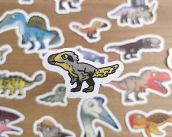 May include: A collection of colorful dinosaur stickers, including a detailed gray and yellow raptor. Other dinosaurs in the assortment include a blue Spinosaurus, a brown T-Rex, and a purple Triceratops. The stickers are on a light brown surface.