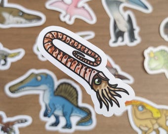 May include: A collection of dinosaur stickers, featuring various species in cartoon style. The stickers include a blue Spinosaurus, a pink Pterodactyl, and a brown and tan ammonite. The stickers are on a wooden surface.