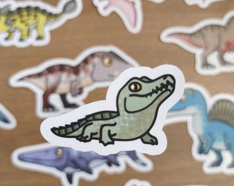 May include: A collection of dinosaur stickers in various colors and designs. The stickers feature different types of dinosaurs, including a green alligator-like dinosaur. The stickers are on a wooden surface.