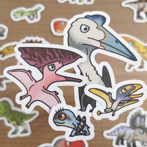 May include: A collection of colorful dinosaur stickers, featuring various species in cartoon style. The stickers include a pink Pterodactyl, a blue and yellow Pterosaur, and a green Tyrannosaurus Rex. The stickers are on a light brown surface.