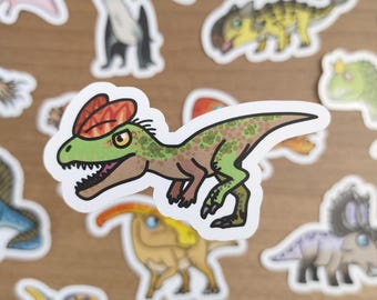 May include: A collection of colorful dinosaur stickers. The stickers feature various dinosaurs in cartoon style, including a Dilophosaurus, Pterodactyl, and Triceratops. The stickers are on a light brown surface.